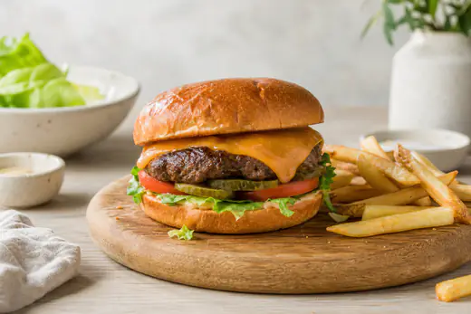Burger wołowy z serem cheddar, sałatą, pomidorem i ogórkiem w bułce, podany na drewnianej desce z frytkami